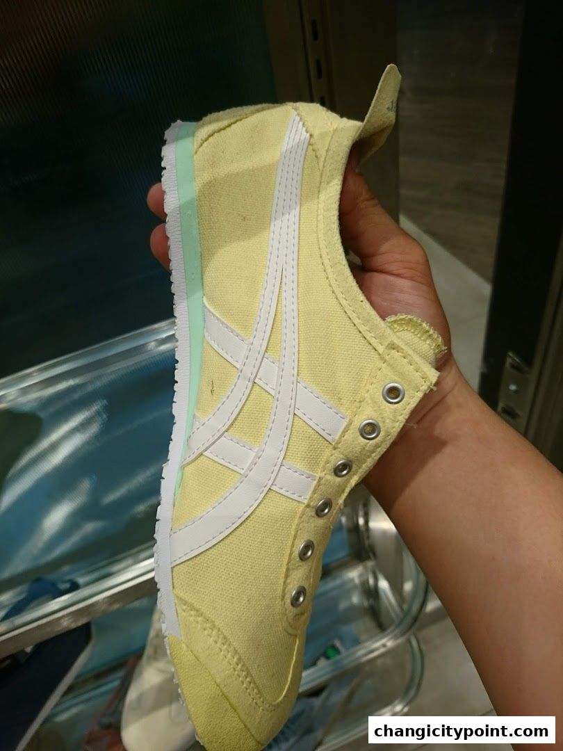 A hand holds a yellow Onitsuka Tiger sneaker with white stripes.