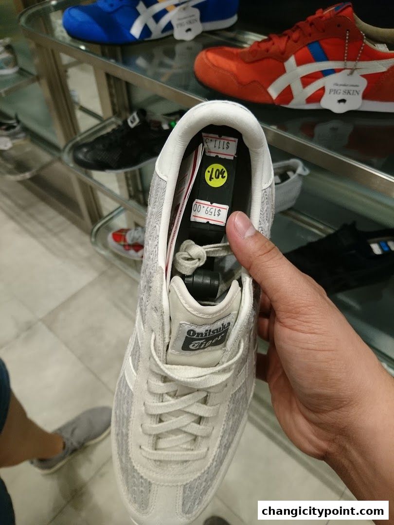 A hand holds a light-colored Onitsuka Tiger sneaker with price tags inside.