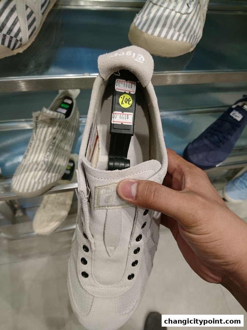 A hand holds a light grey Onitsuka Tiger sneaker with price tags attached.