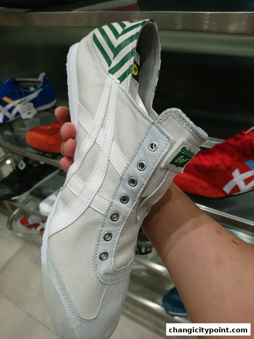 A hand holds a white Onitsuka Tiger shoe with green accents against a backdrop of other shoes.