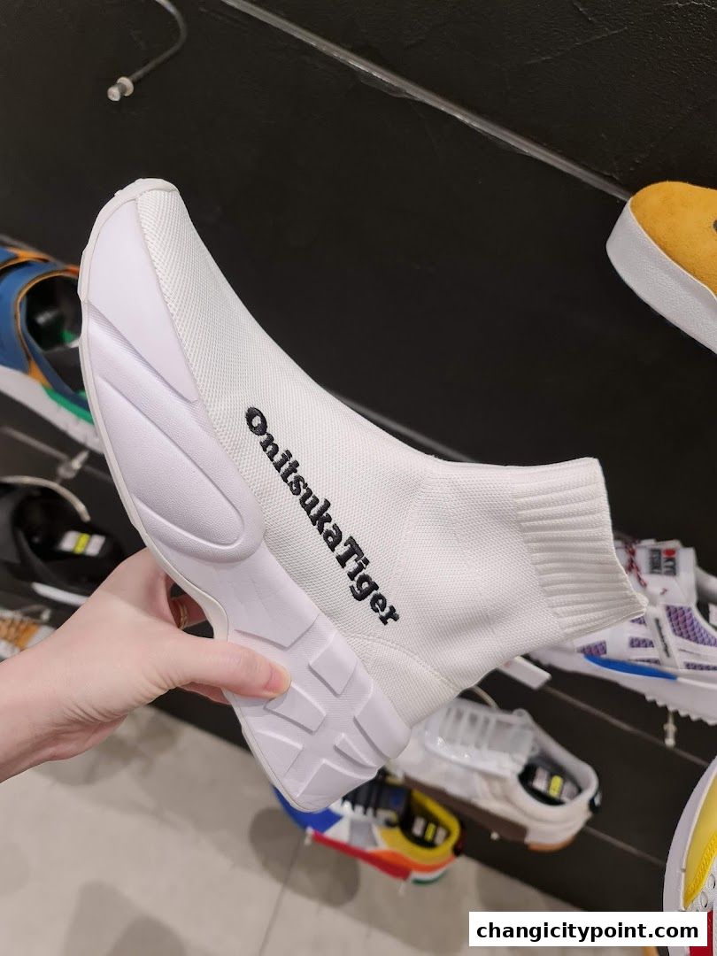 A hand holds a white Onitsuka Tiger sock-style sneaker with black logo.