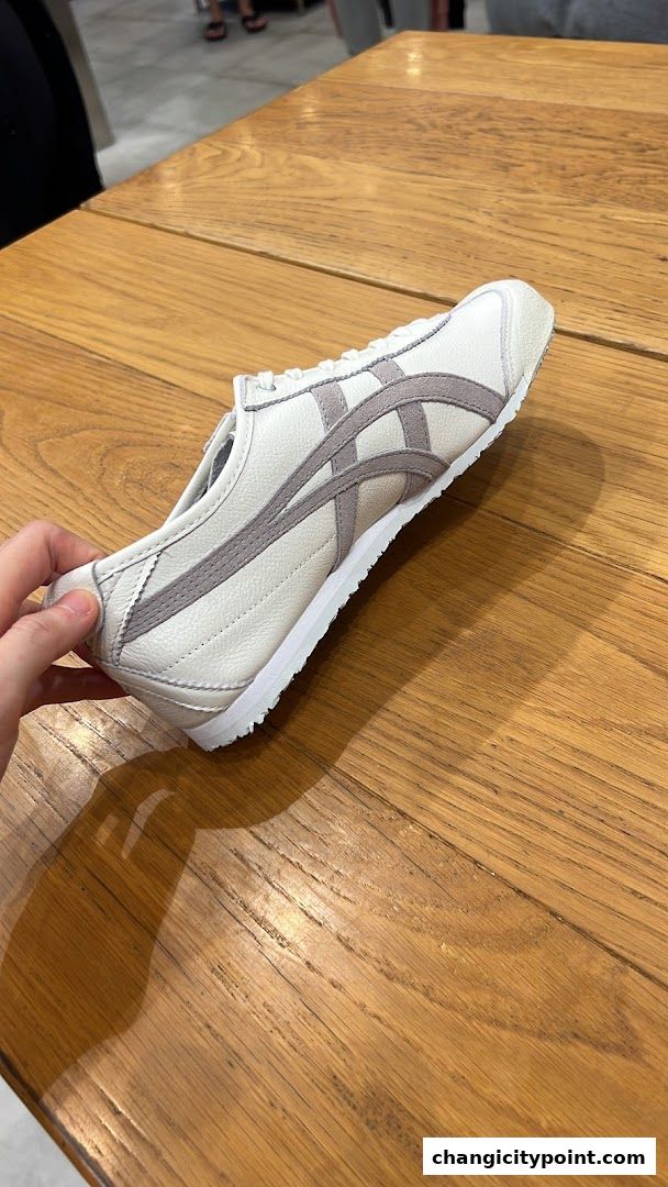 A hand holds a white Onitsuka Tiger sneaker with grey stripes on a wooden table.