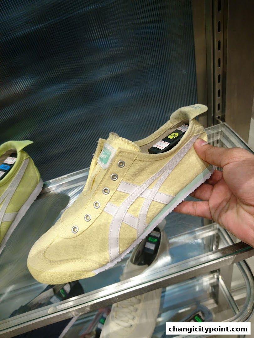 A hand holds a light yellow Onitsuka Tiger sneaker with white stripes.