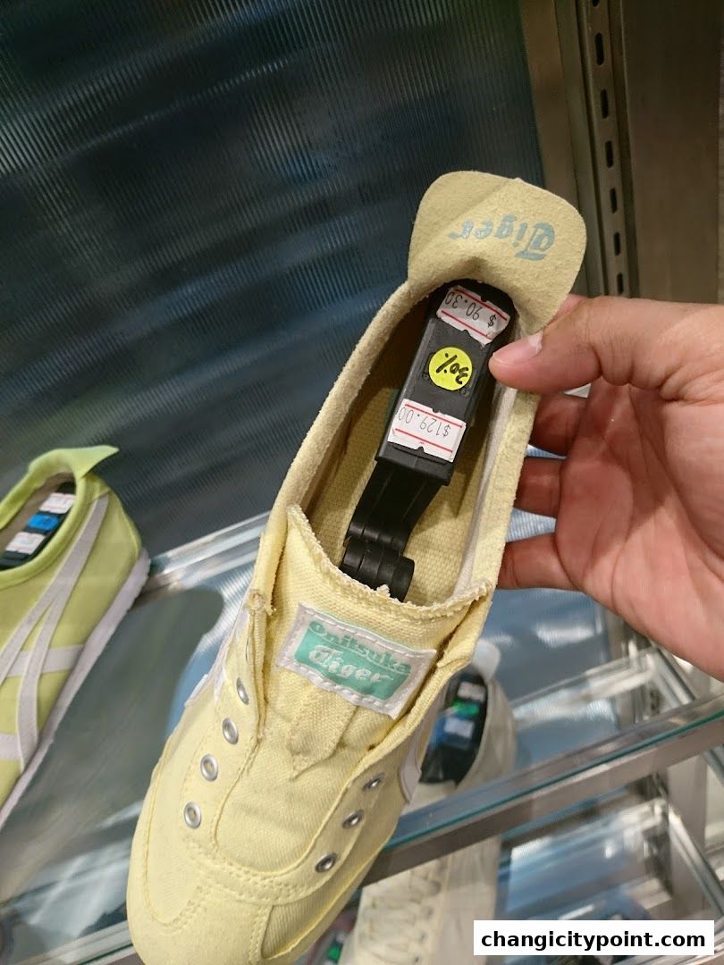A hand holds a yellow Onitsuka Tiger shoe with a price tag showing a discount.
