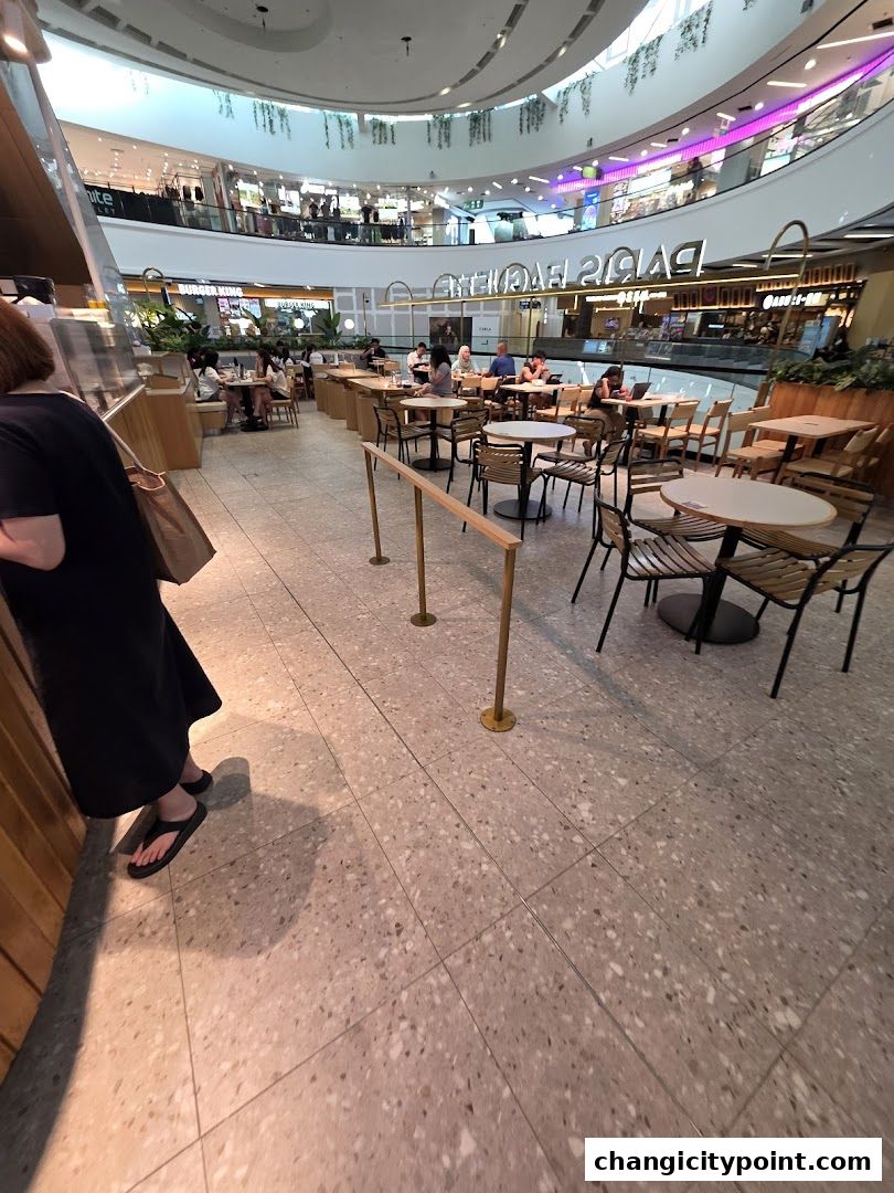 Interior view of Paris Baguette cafe with seating areas and mall shops in the background.