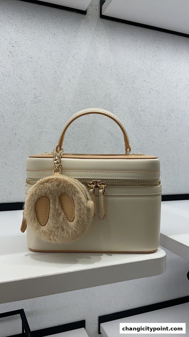 A cream-colored structured handbag with a fluffy charm is displayed on a shelf.
