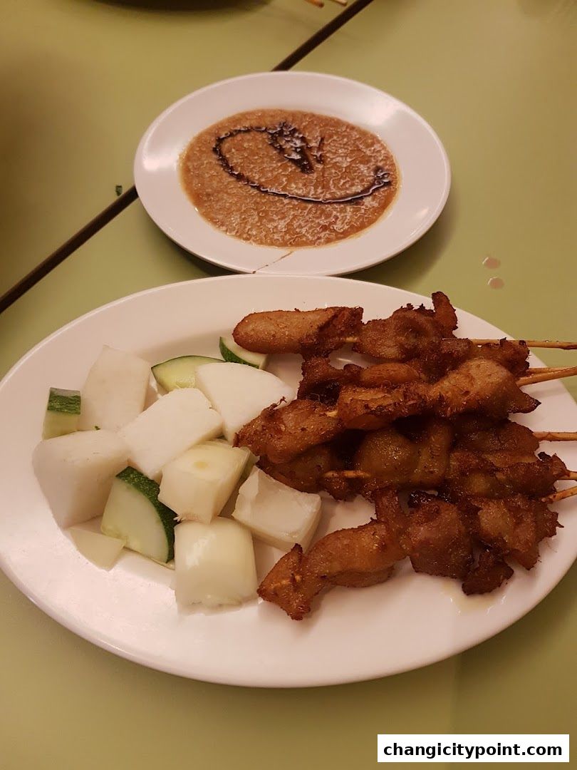 A plate of satay with peanut sauce and rice cakes, served with cucumber and onion.