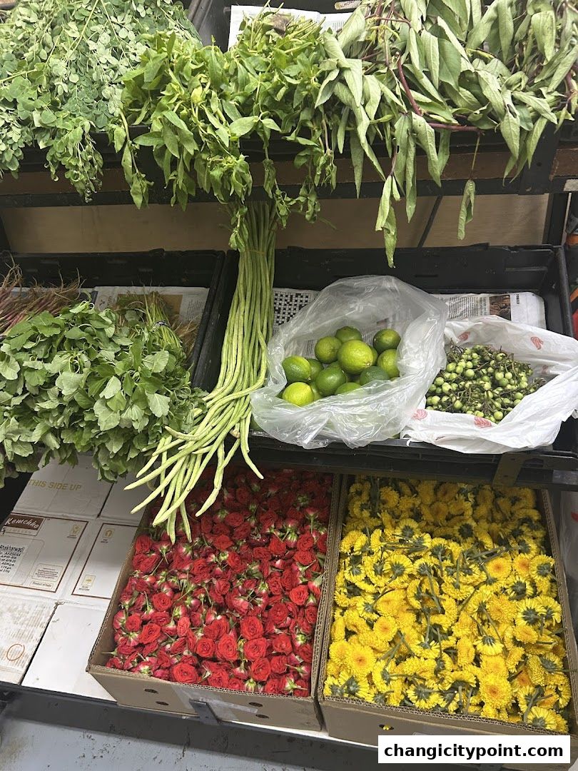 A display of fresh produce including long beans, limes, herbs, red roses, and yellow flowers.