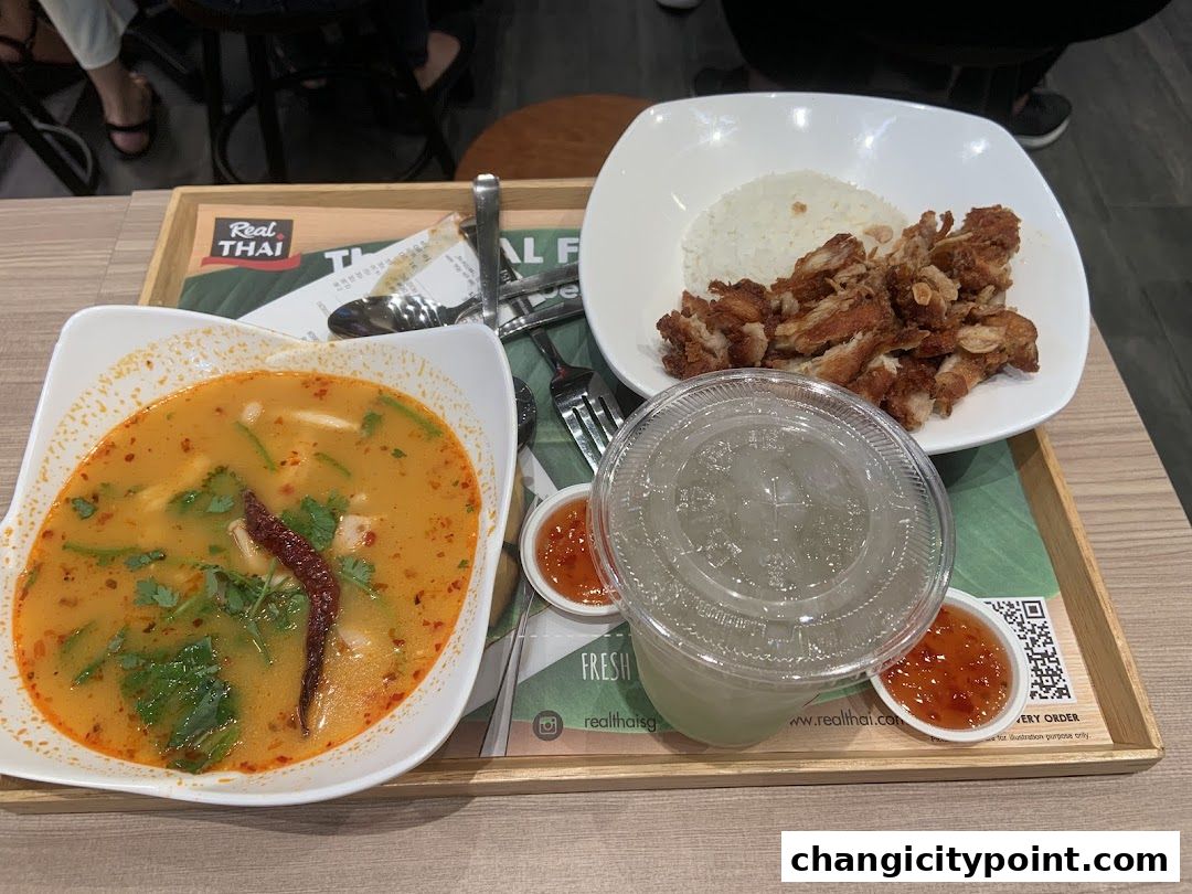 A tray with Thai food including Tom Yum soup, fried chicken with rice, and a drink.