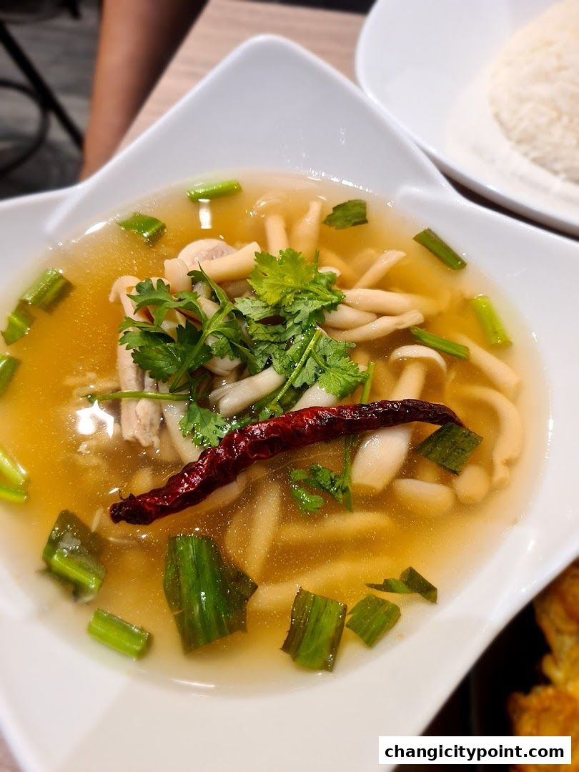 A close-up of a steaming bowl of Thai soup with mushrooms, chicken, and chili.