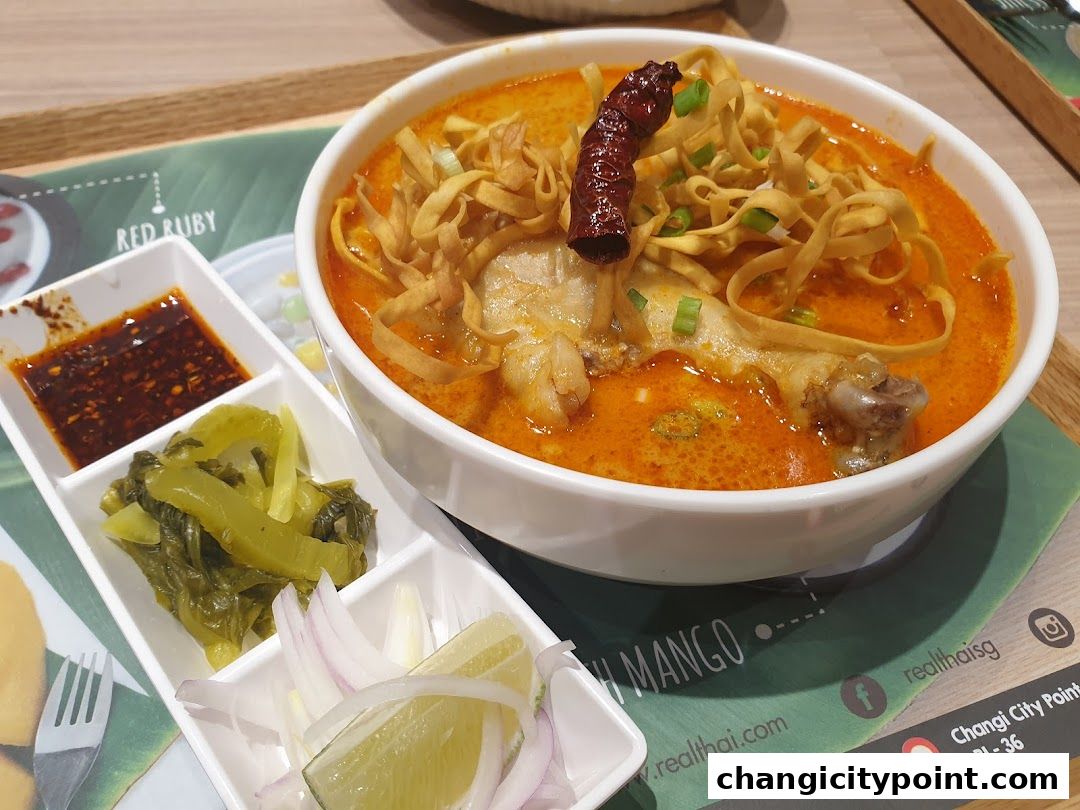 A bowl of Khao Soi curry noodle soup with chicken, served with pickled vegetables, chili paste, and lime.