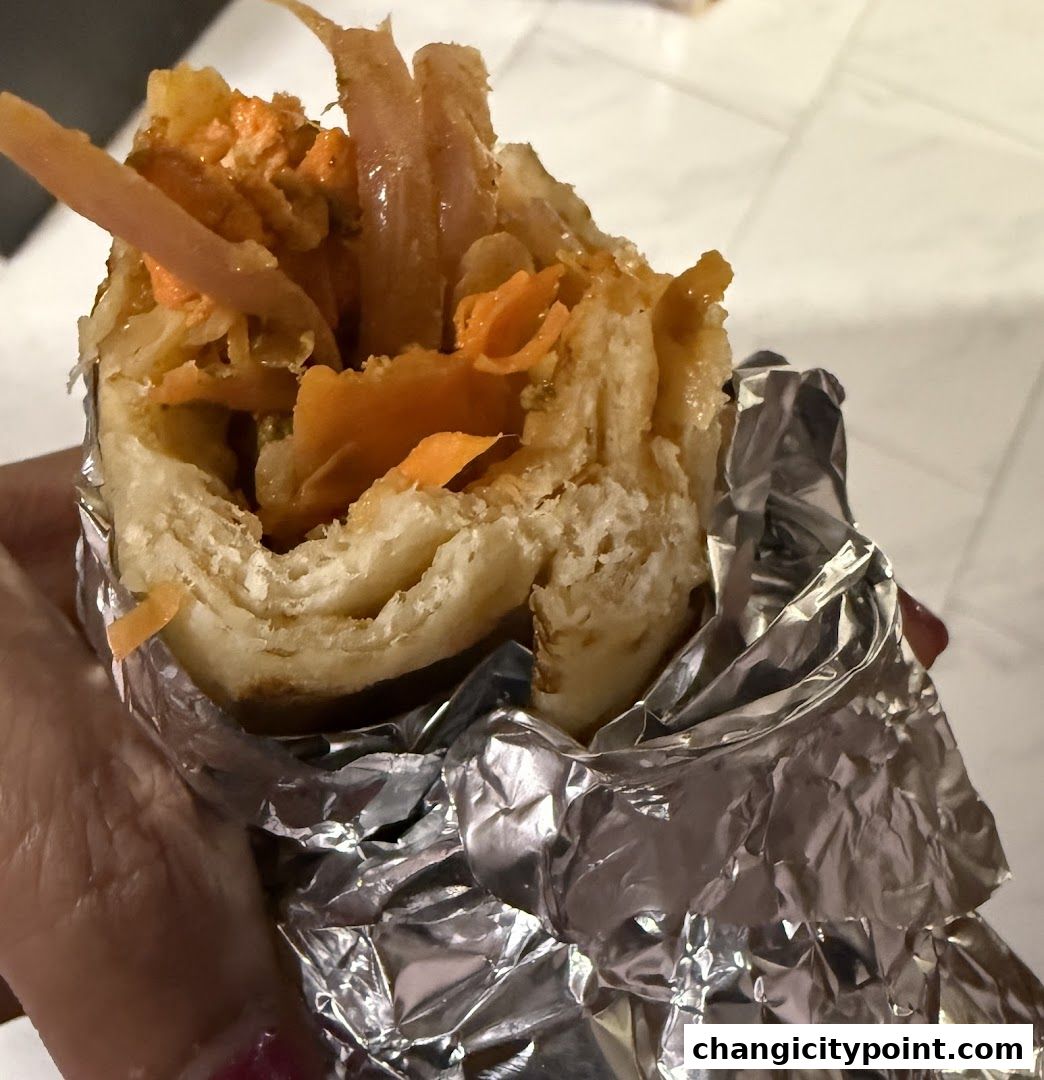 A hand holds a delicious-looking roll wrapped in foil, filled with meat and vegetables.