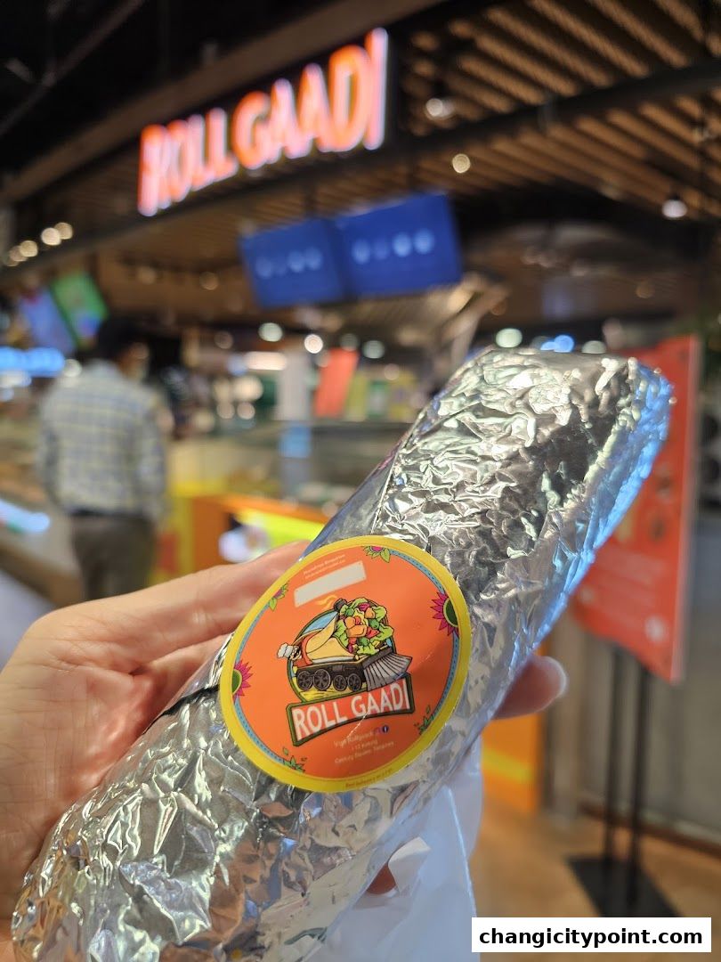 A hand holds a foil-wrapped roll with the RollGaadi logo, with the shop's signage visible in the background.