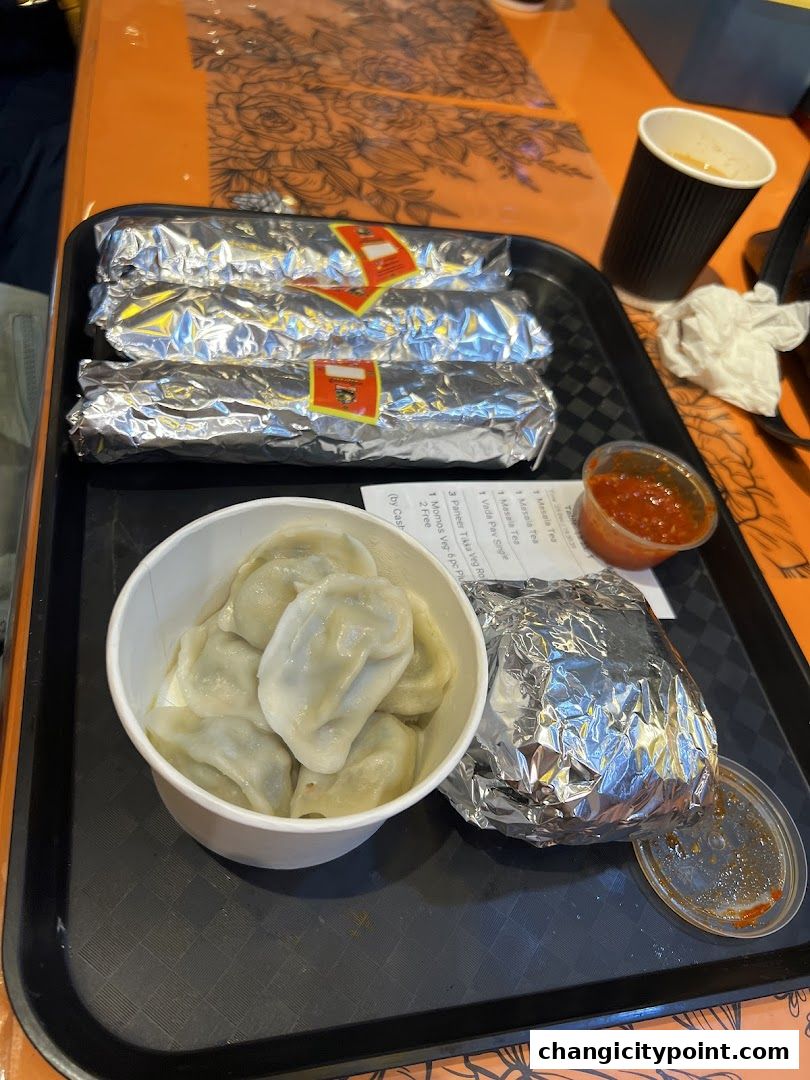 A tray with momos, foil-wrapped items, dipping sauce, and a cup of tea.