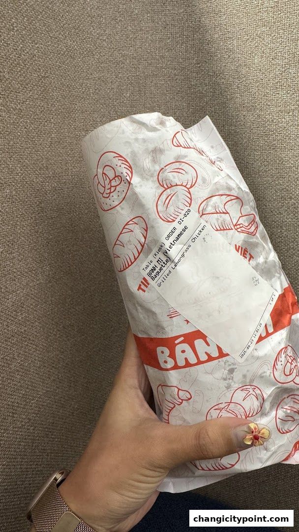 A hand holds a wrapped Vietnamese Banh Mi sandwich with grilled lemongrass chicken.