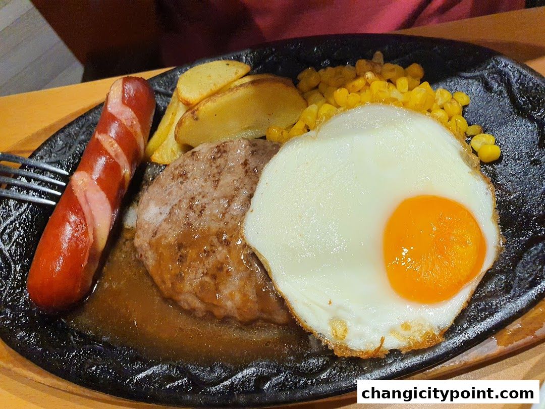 A sizzling plate of food featuring a hamburger steak, sausage, fried egg, corn, and potato wedges.