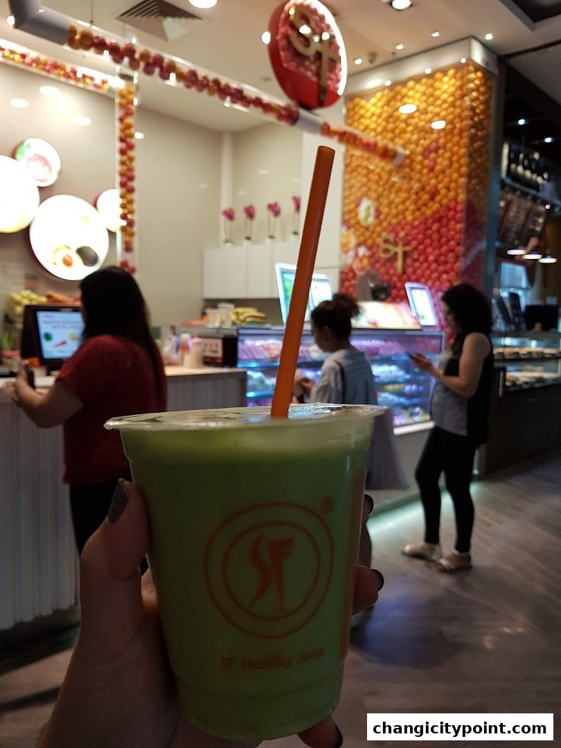 A hand holds a green juice in front of a brightly decorated fruit juice shop.