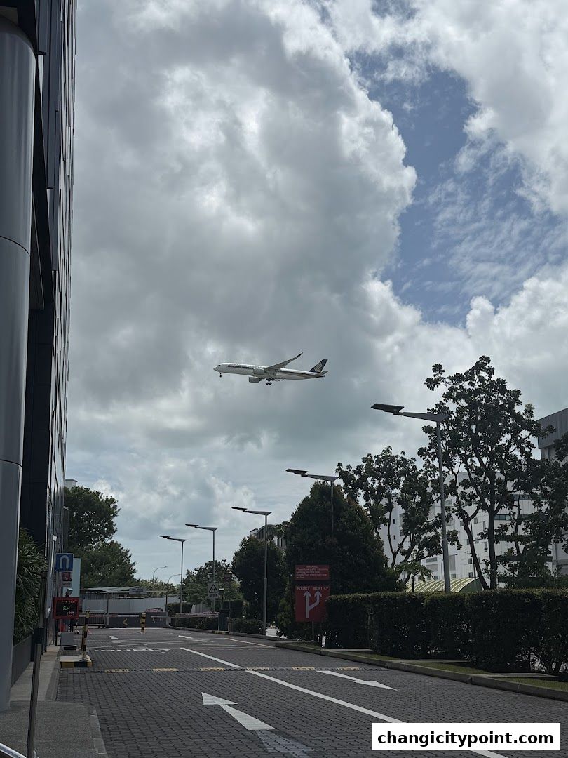 An airplane flies over a road with trees and buildings under a cloudy sky.
