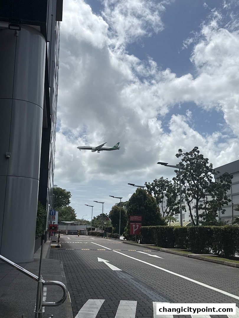 An airplane flies over a street with a building on the left.