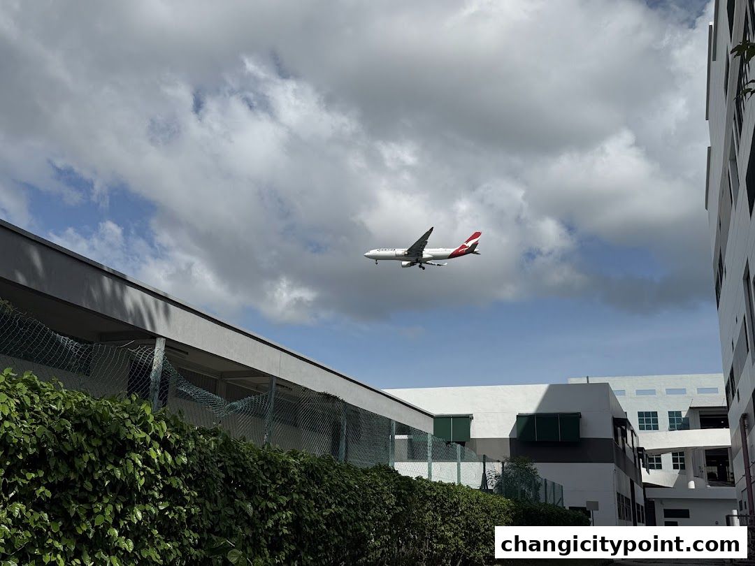 An airplane flies over buildings and lush greenery under a cloudy sky.