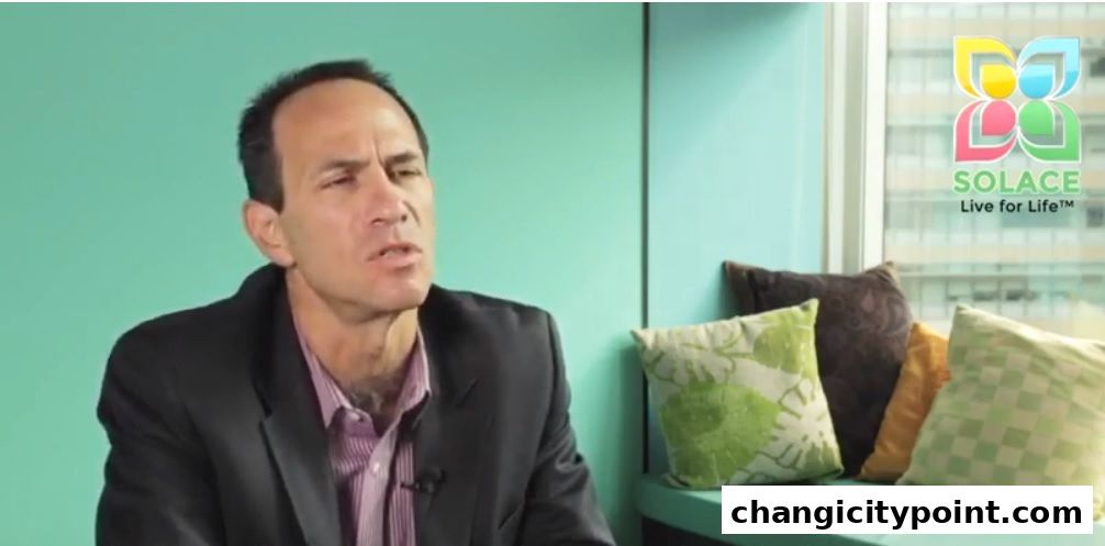 A man in a suit sits in front of a teal wall with decorative pillows.