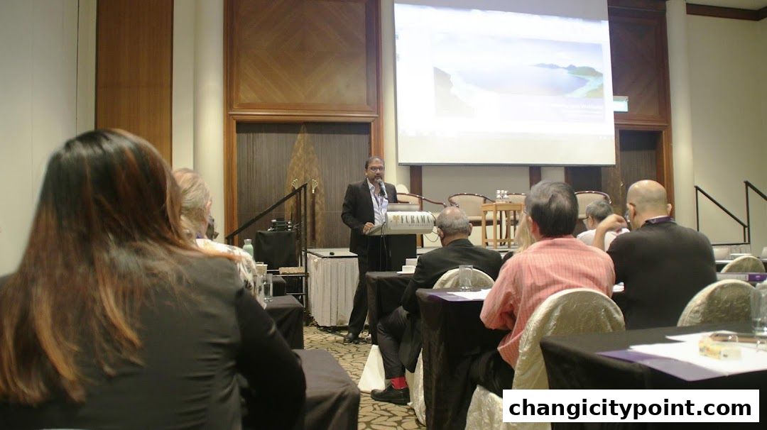 A speaker presents to an audience in a conference room with a projector screen.