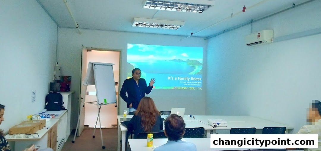 A man presents a lecture titled 'It's a Family Illness' to an audience.