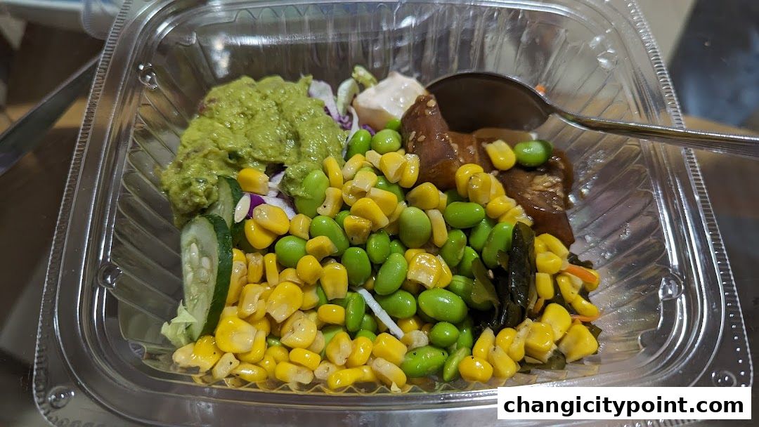 A clear plastic container filled with a colorful salad featuring corn, edamame, guacamole, and cucumber.