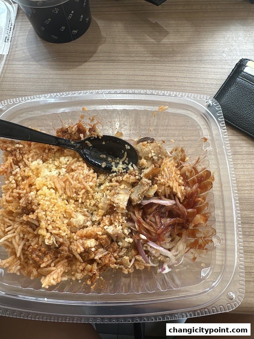 A take-out container filled with rice, meat, and toppings, with a spoon and wallet nearby.