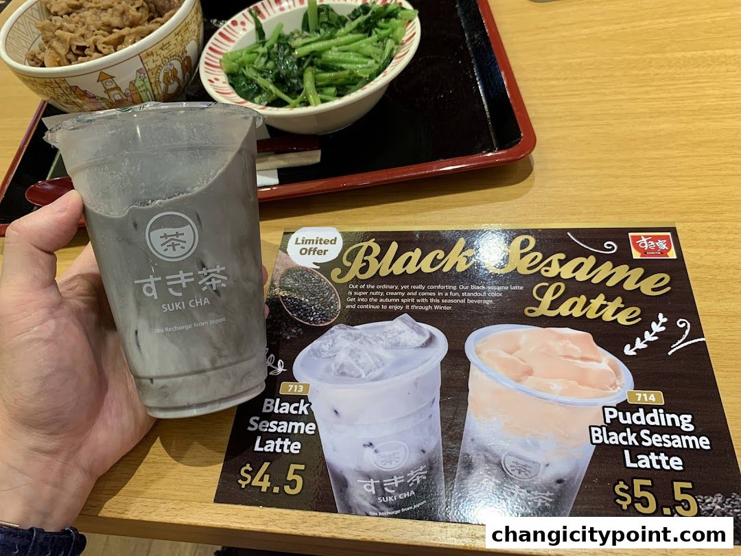 A hand holds a black sesame latte next to a menu featuring black sesame drinks.