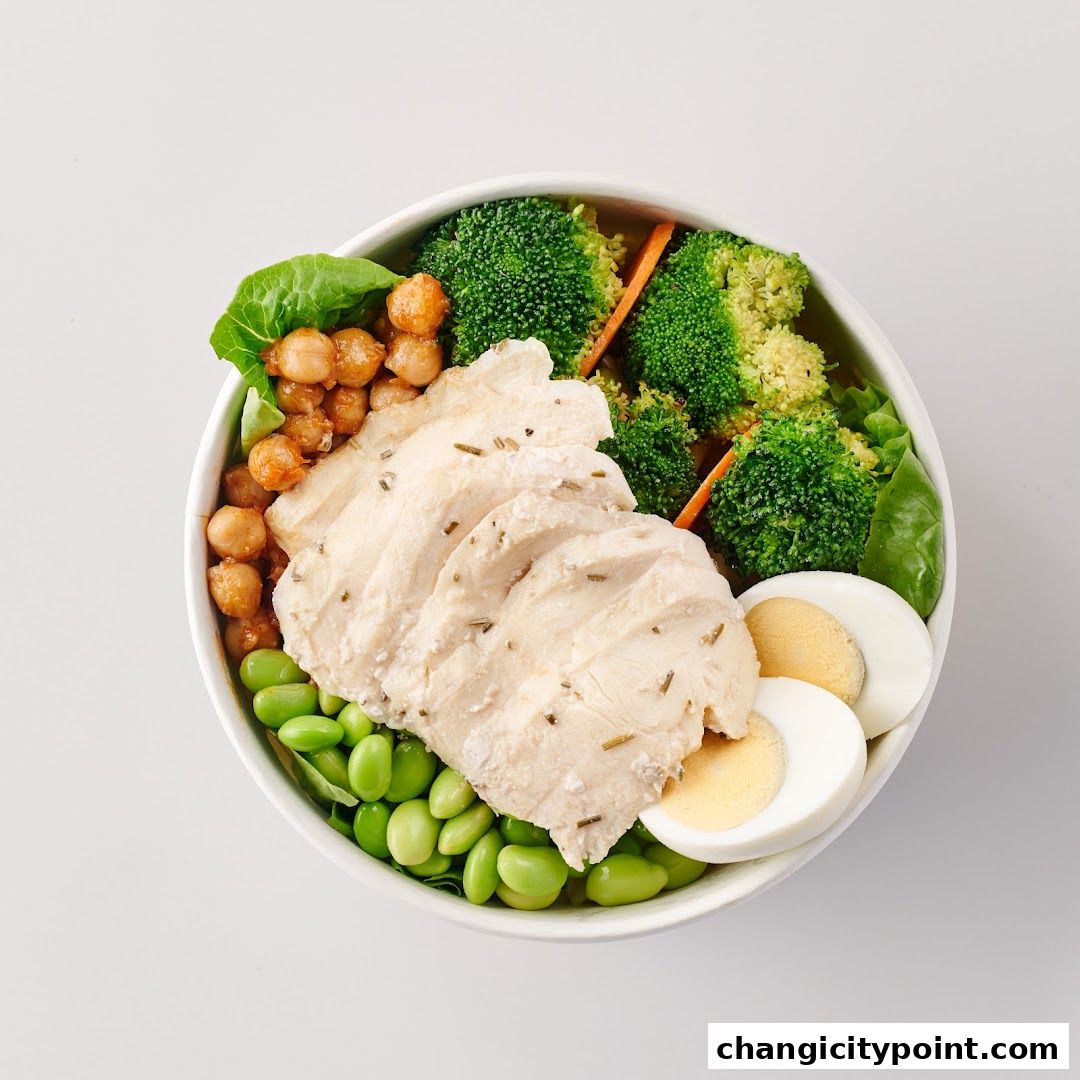 A healthy bowl of sliced chicken breast, edamame, chickpeas, broccoli, and egg.
