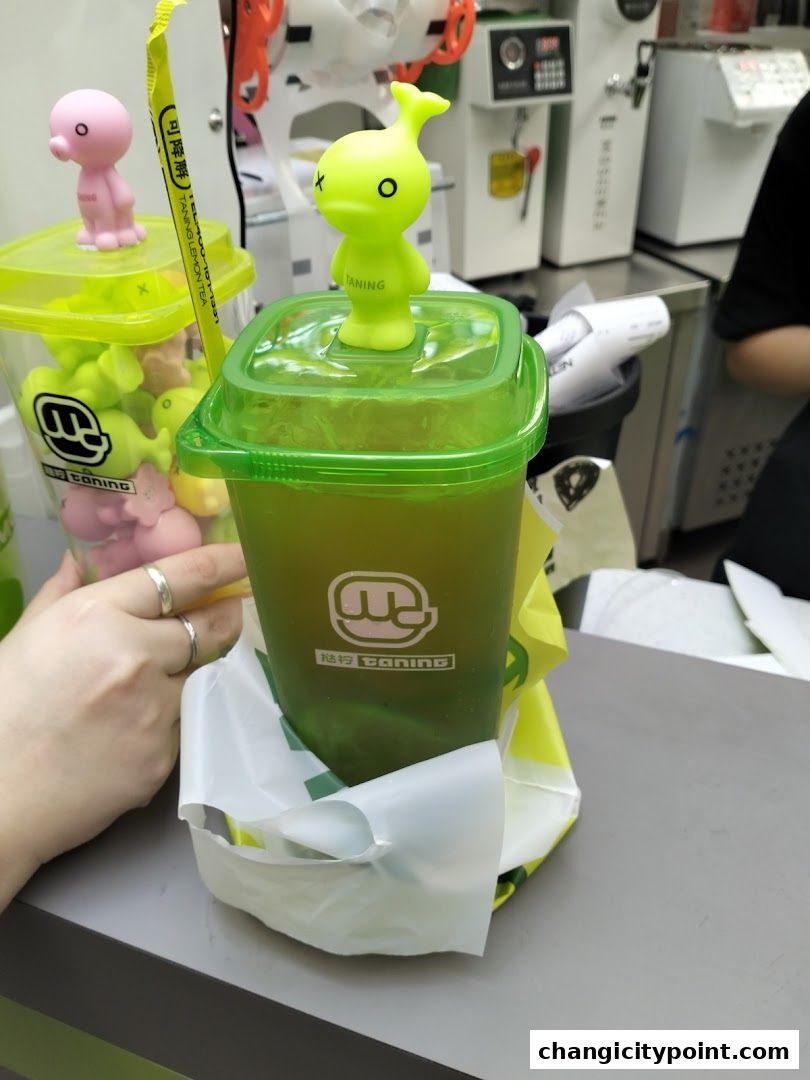 A green beverage with a cute toy on top, in a plastic cup with a shopping bag.