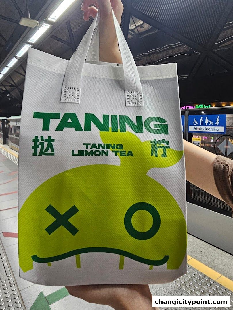 A person holds a white tote bag with "TANING LEMON TEA" branding and a cartoon character.