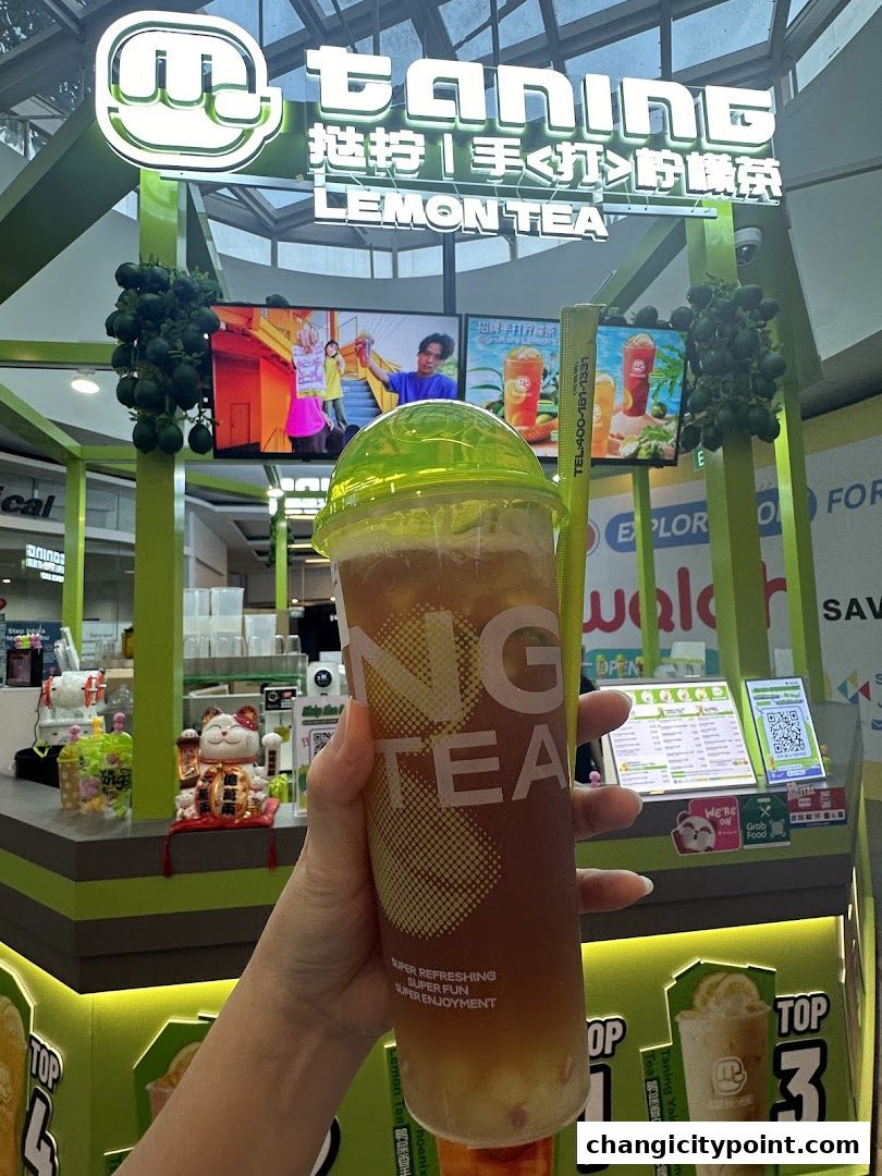 A hand holds a refreshing lemon tea drink in front of the Taning Lemon Tea shop.