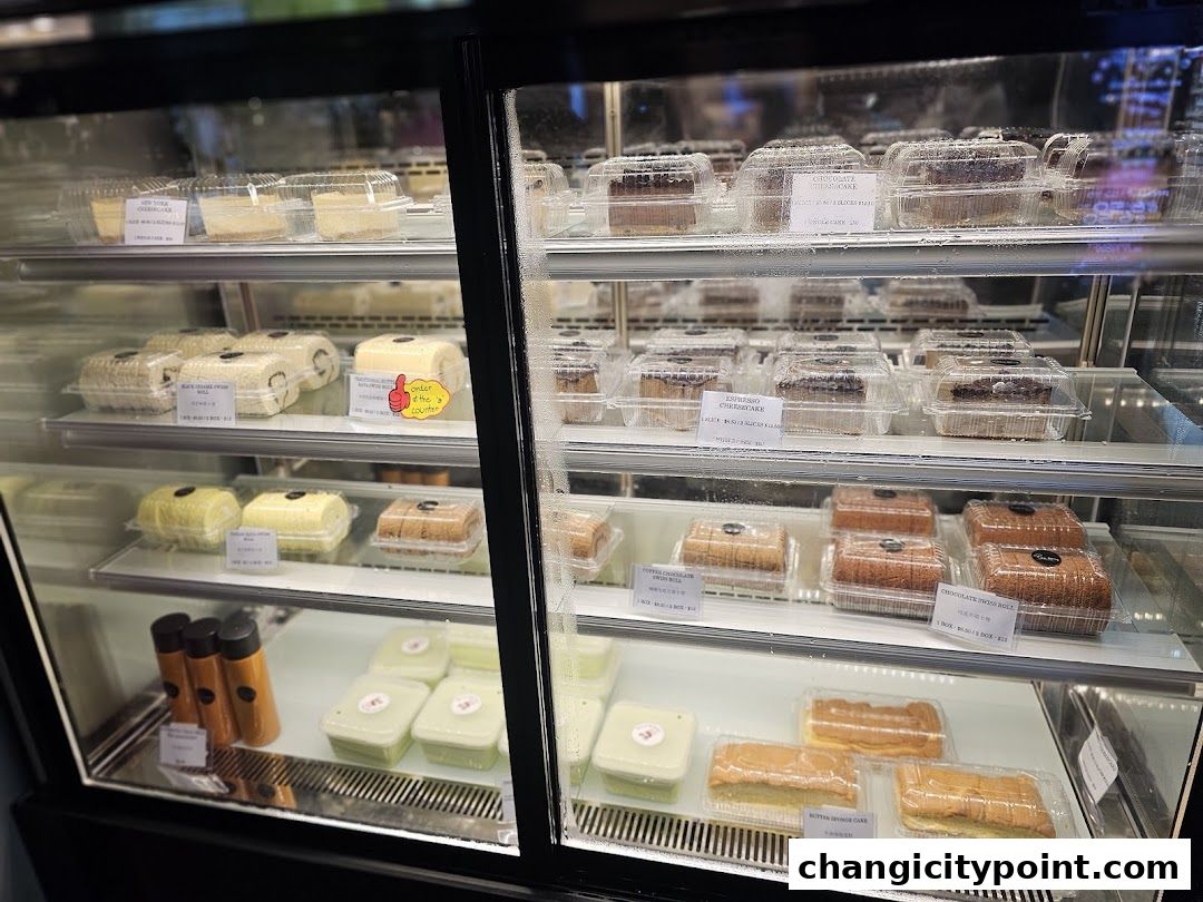 A display fridge filled with various cakes, swiss rolls, and bottled drinks.