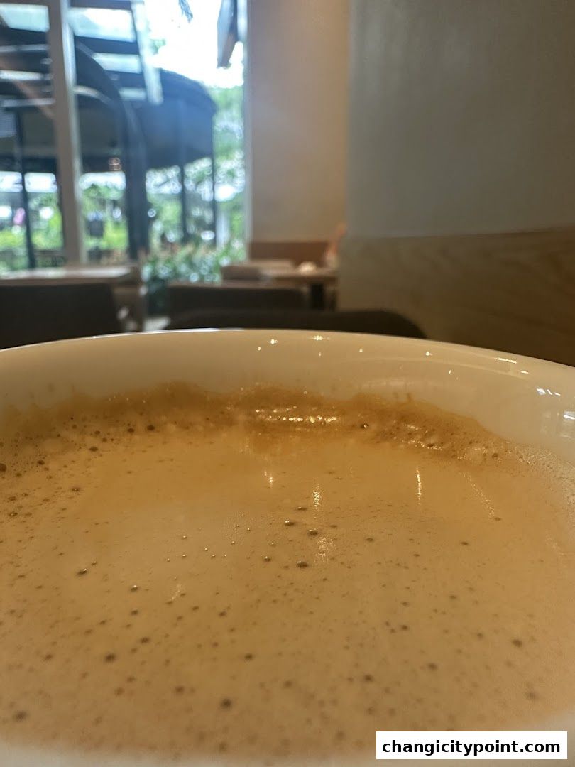 A close-up of a frothy coffee drink in a white mug, with a blurred background of a cafe.