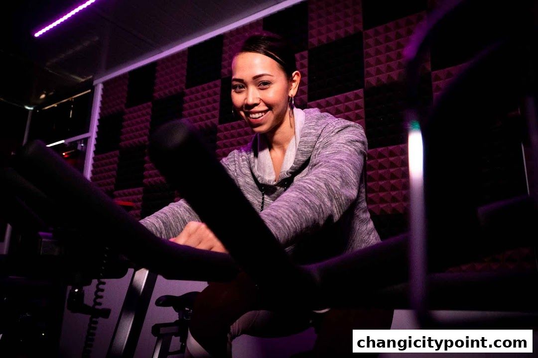 A smiling woman rides an exercise bike in a dimly lit gym with purple lighting.