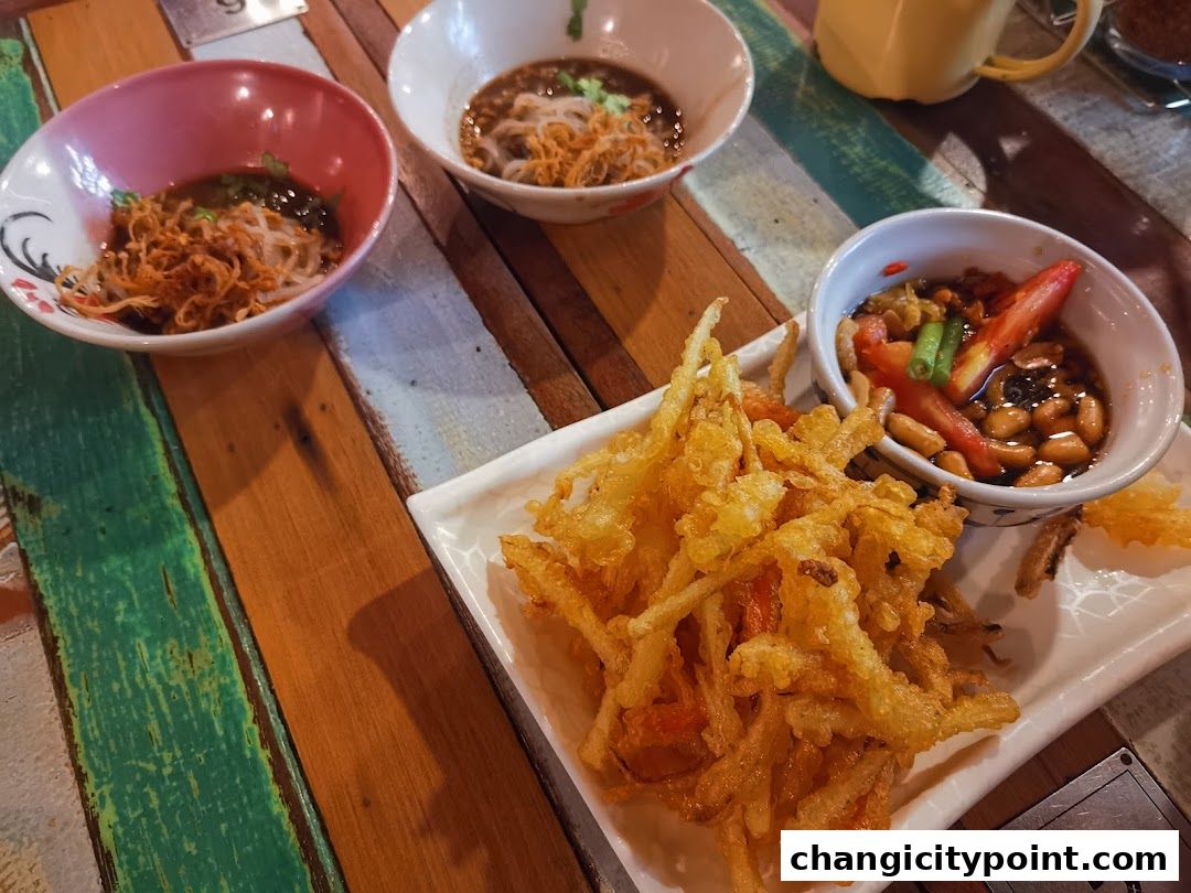 A close-up of delicious Thai boat noodles and crispy fried snacks served on a colorful wooden table.