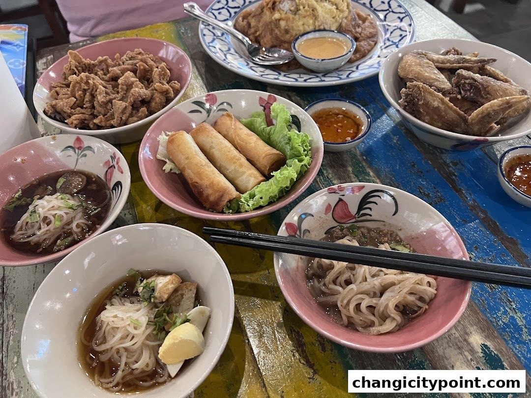 A table laden with various Thai dishes including noodles, spring rolls, fried chicken, and wings.