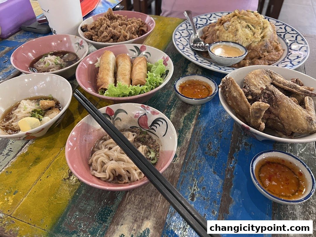 A table laden with various Thai dishes including noodles, spring rolls, fried chicken, and omelets.