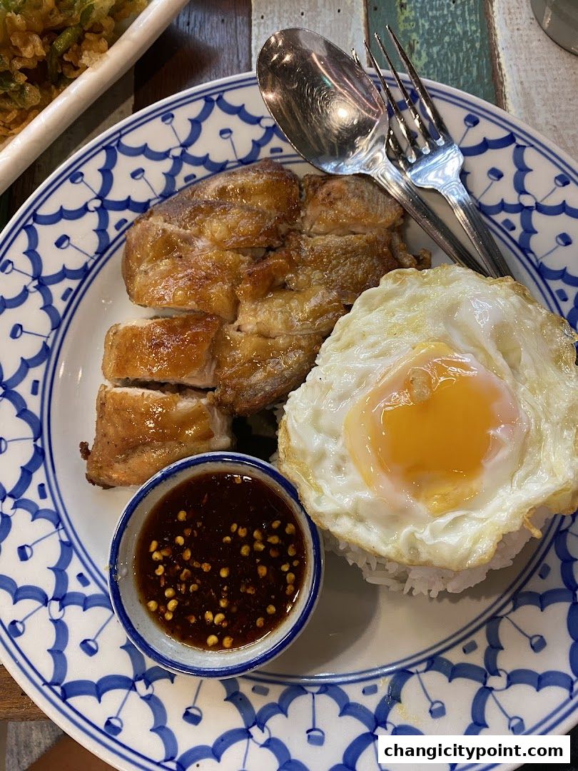 A plate of rice topped with grilled chicken, a fried egg, and a side of chili sauce.