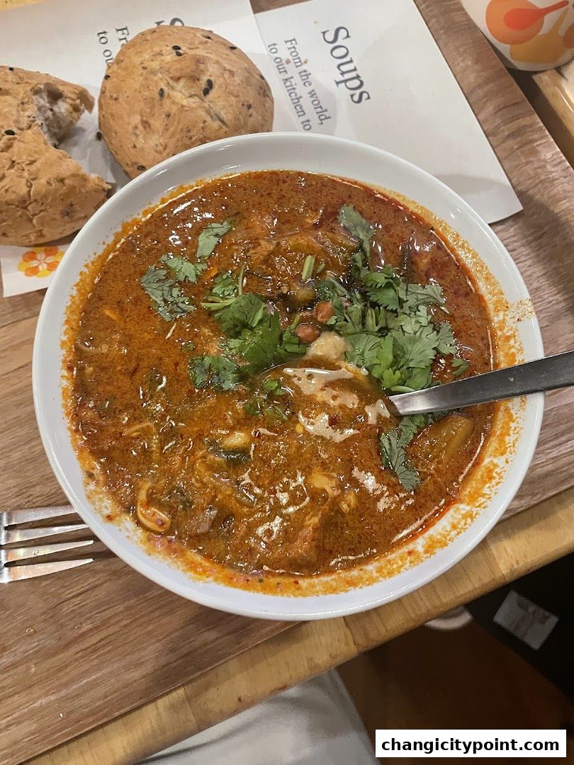 A bowl of rich, spicy soup with fresh cilantro and a side of bread rolls.