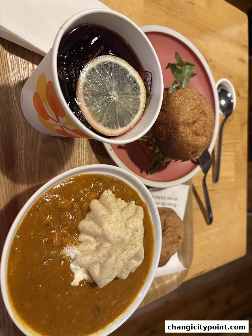A bowl of soup with a cracker, a small bun, and a drink with lemon.