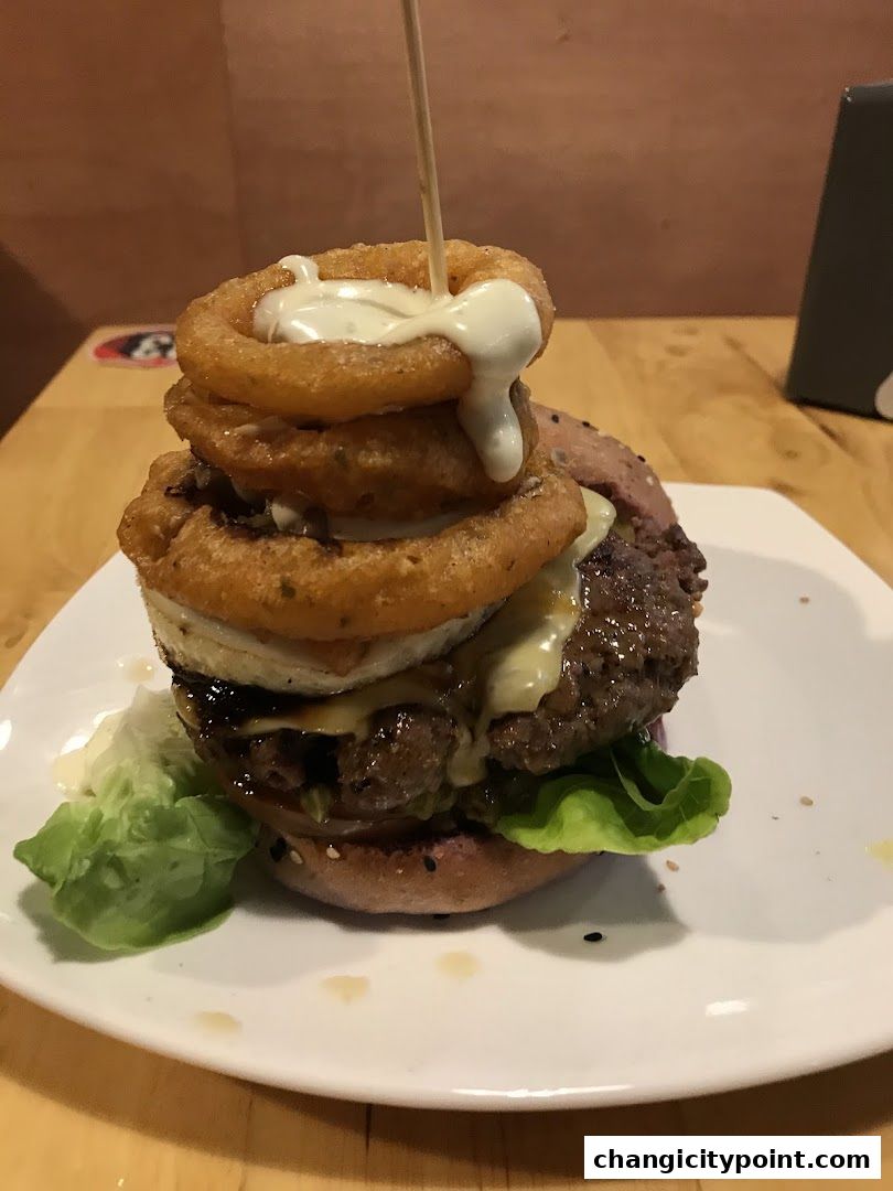 A tall burger with onion rings and sauce on a white plate.