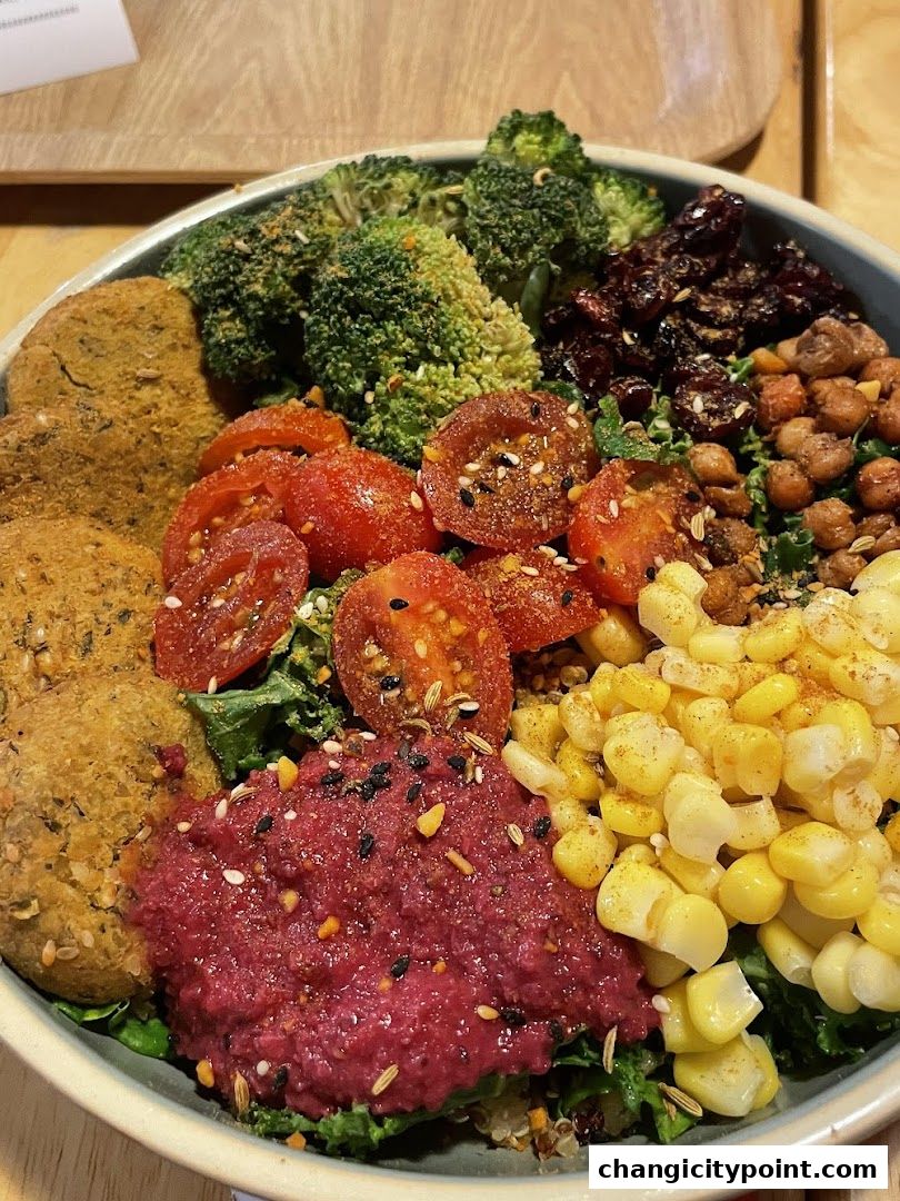 A vibrant and healthy bowl filled with broccoli, tomatoes, corn, falafel, and beet hummus.