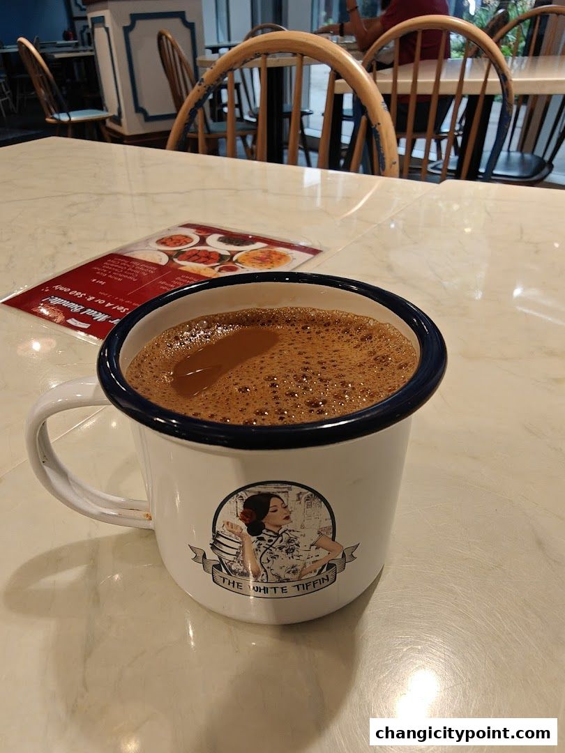 A close-up of a frothy beverage in a mug with The White Tiffin logo.