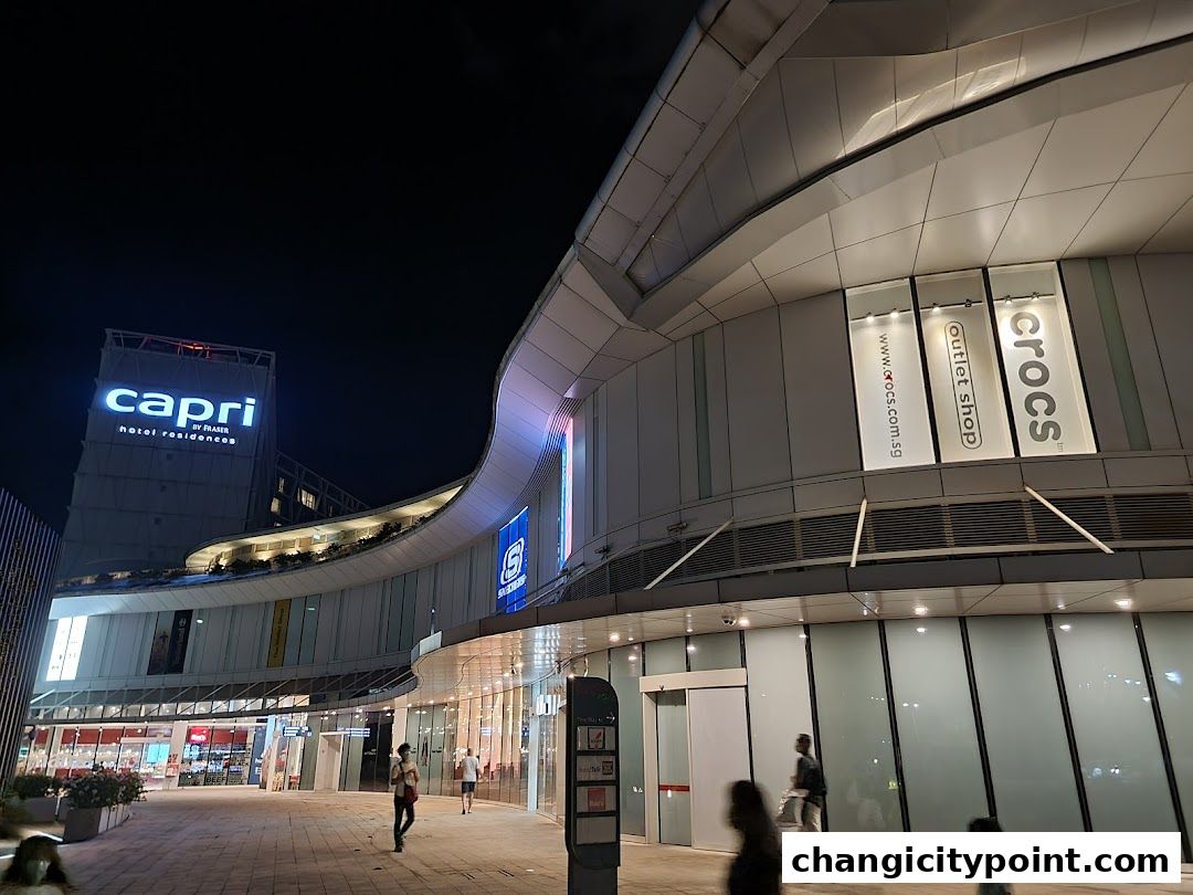 Exterior view of a shopping mall at night, featuring the Capri by Fraser hotel and various shops.