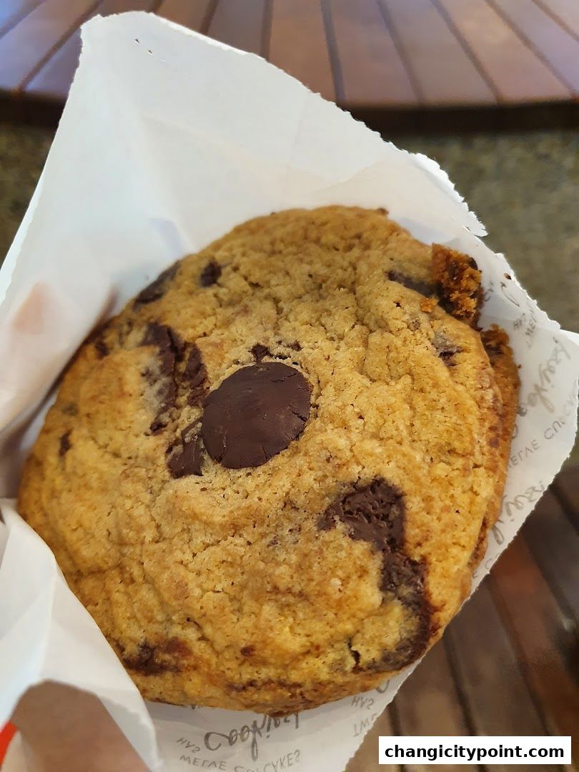 A close-up shot of a freshly baked chocolate chip cookie in a white paper bag.