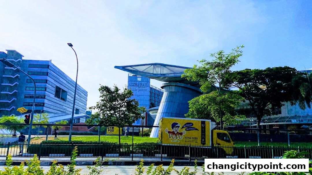 A yellow delivery truck parked in front of modern buildings and lush greenery.