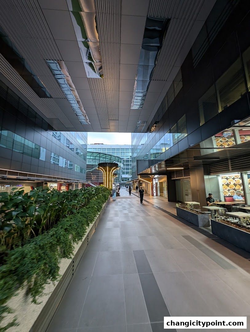 A modern outdoor walkway with shops and greenery, leading towards a bright archway.